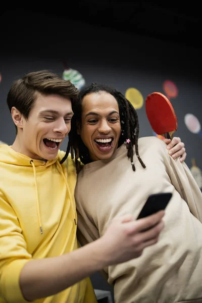 Amis interraciaux positifs avec raquette de tennis en utilisant le téléphone mobile dans le club de jeu — Photo de stock