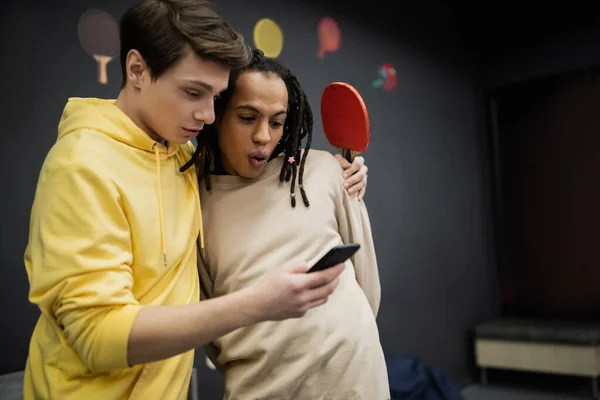 Jeune homme utilisant un smartphone et tenant raquette de tennis près choqué ami multiracial dans le club de jeu — Photo de stock