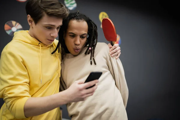 Amigos multiétnicos conmocionados con raqueta de tenis usando teléfono inteligente en el club de juegos - foto de stock