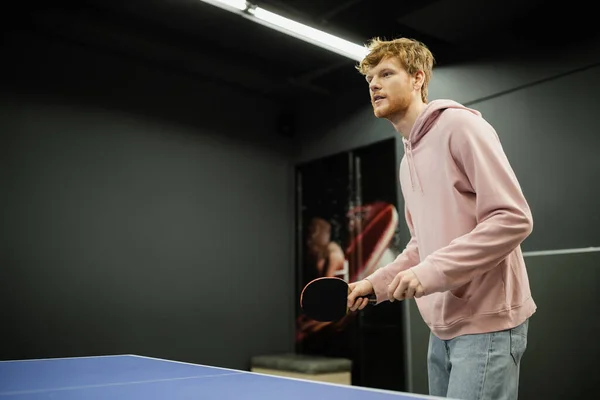 Joven pelirroja sosteniendo raqueta mientras juega al tenis de mesa en el club de juegos - foto de stock