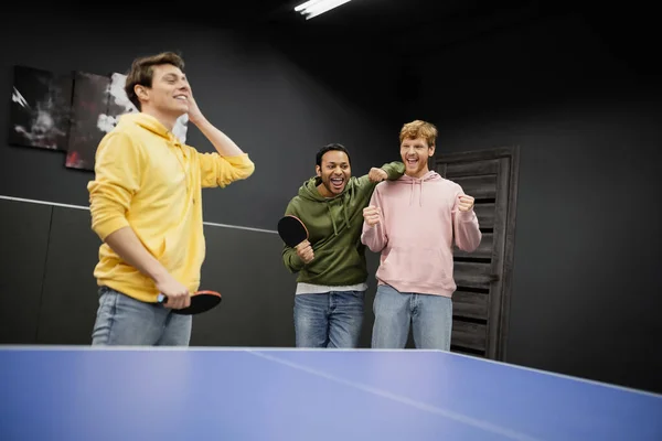 Excited interracial men standing near blurred friend with tennis racket in gaming club — Stock Photo