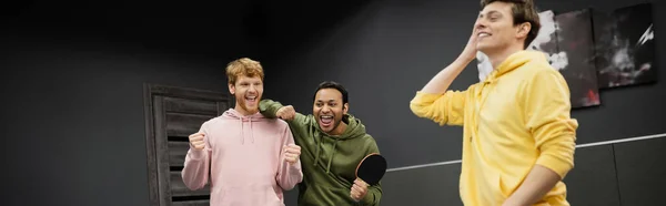 Fröhliche gemischtrassige Männer mit Tennisschläger zeigen Ja-Geste nahe Freund in Spielklub, Banner — Stockfoto