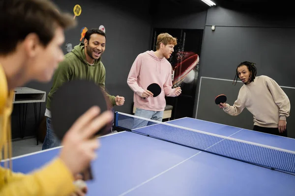 Возбужденные межрасовые мужчины, стоящие рядом с друзьями, играют в настольный теннис в игорном клубе — стоковое фото