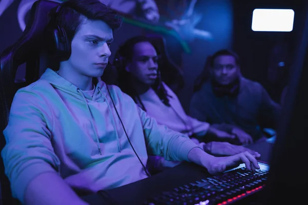 Hombre enfocado en auriculares jugando videojuegos cerca de amigos difusos multiétnicos en el club cibernético - foto de stock