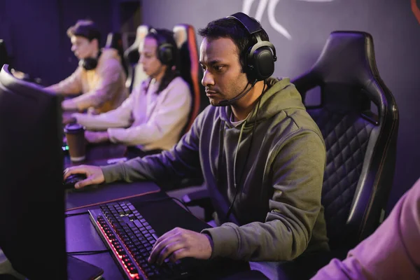 Индийский геймер в наушниках играет в видеоигры на компьютере рядом с размытой командой в игорном клубе — стоковое фото