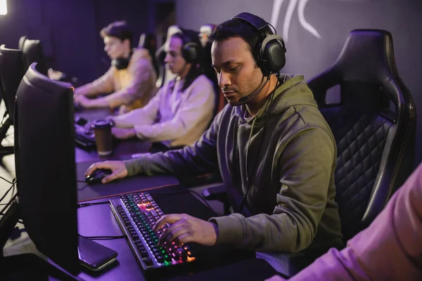 Indischer Gamer mit Kopfhörer und Mikrofon spielt Videospiel auf Computer in der Nähe verschwommener Freunde in Spielclub — Stockfoto