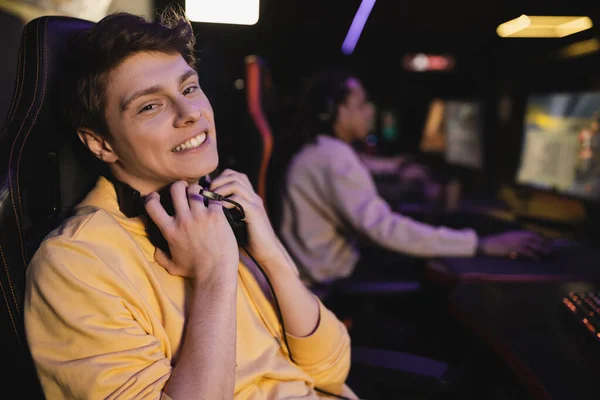 Carefree young man holding headphones and looking at camera in cyber club — Stock Photo