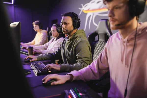 Indischer Spieler mit Kopfhörern spielt Videospiel auf Computer in der Nähe verschwommenes Team in Cyber-Club — Stockfoto