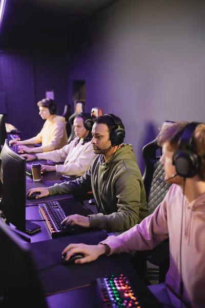 Indischer Spieler mit Kopfhörer spielt Videospiel auf Computer in der Nähe verschwommener Freunde in Spielclub — Stockfoto