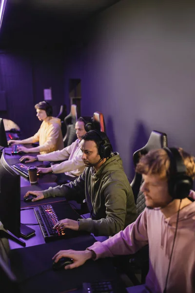 Indischer Spieler mit Kopfhörer und Mikrofon spielt Videospiel am Computer in der Nähe von Team im Cyber-Club — Stockfoto