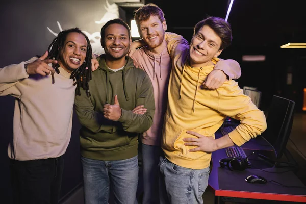 Cheerful interracial friends gesturing and looking at camera in gaming club — Stock Photo