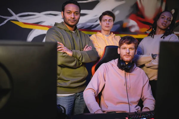 Jogadores multiétnicos olhando para a câmera perto de computadores no clube de jogos — Fotografia de Stock