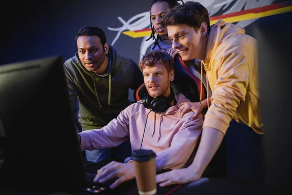 Cheerful multiethnic men looking at friend playing video game on computer in gaming club — Stock Photo