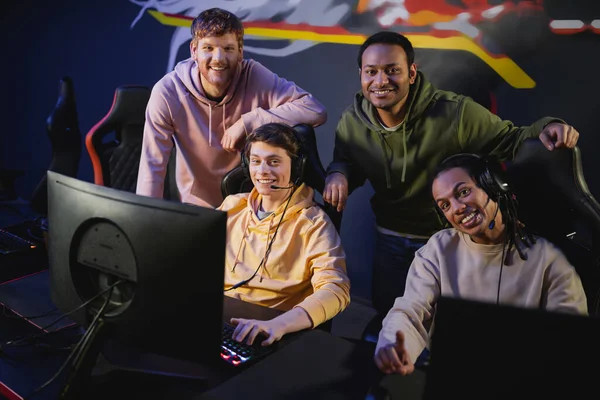 Positive multicultural friends in headphones looking at camera near computers in gaming club — Stock Photo