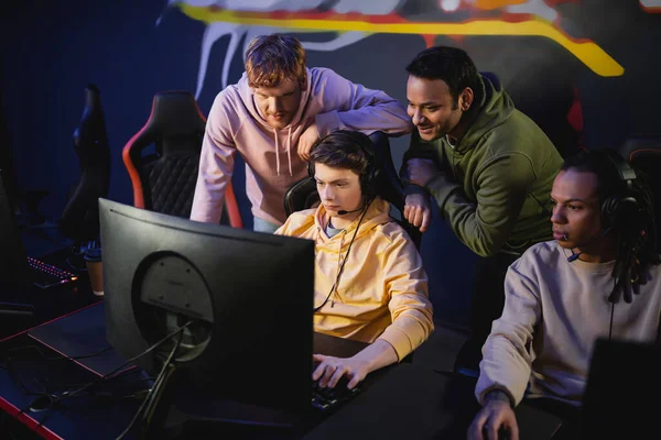 Smiling multiethnic men looking at friend playing video game in gaming club — Stock Photo