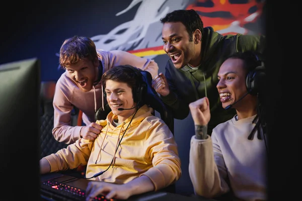 Emocionados hombres interracial mirando a un amigo jugando videojuegos en la computadora en el club de juegos - foto de stock