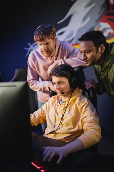 Hombre sonriente en auriculares jugando videojuegos cerca de amigos multiétnicos en el club de juegos - foto de stock