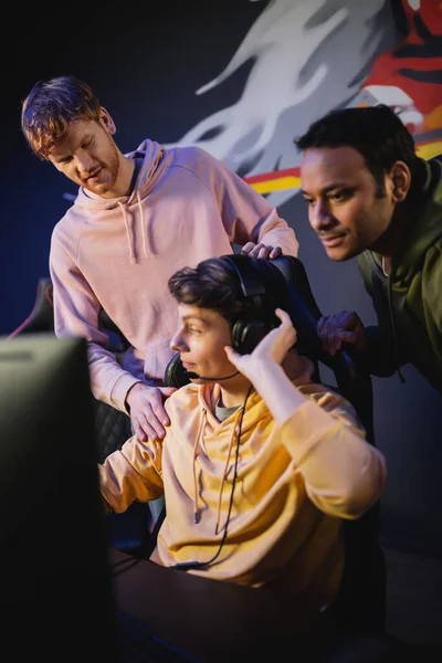 Hombre joven con auriculares cerca de amigos interracial y la computadora en el club de juego - foto de stock