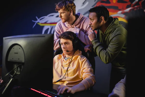 Interracial team standing near gamer playing video game on computer in cyber club — Stock Photo