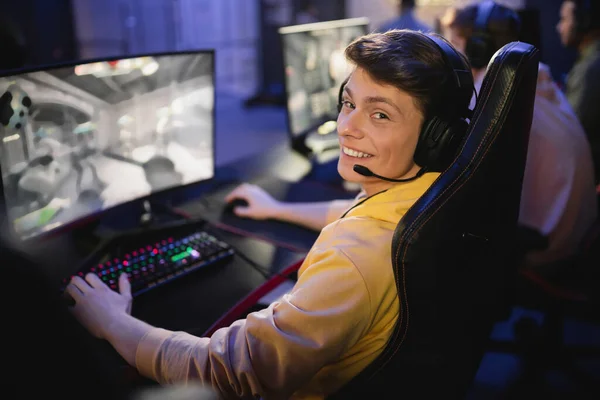 Homem sorridente em fones de ouvido olhando para a câmera enquanto joga vídeo game no clube cibernético — Fotografia de Stock