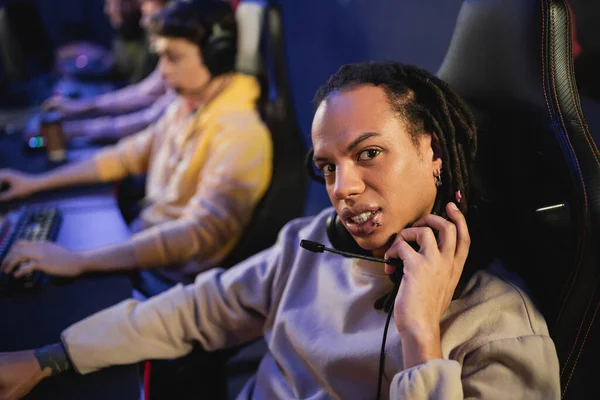 Multiracial gamer holding headphones and looking at camera near blurred team in gaming club — Stock Photo