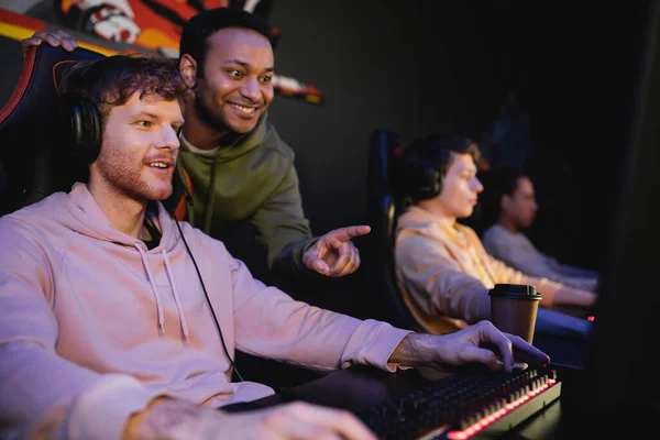 Hombre indio sonriente apuntando a la computadora cerca de un amigo jugando videojuego en el club de juegos - foto de stock