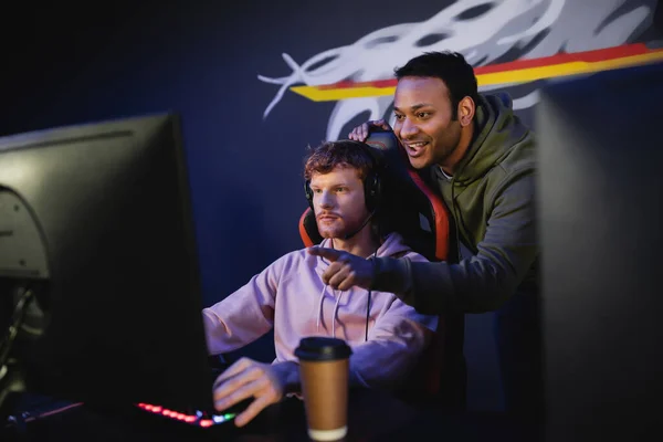 Positive indian man pointing with finger while friend playing video game on computer in gamming club — Stock Photo