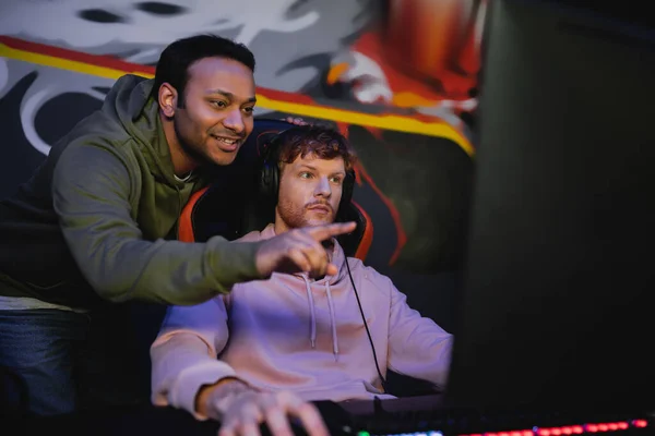 Cheerful indian man pointing at blurred computer monitor while friend in headphones playing video game in cyber club — Stock Photo