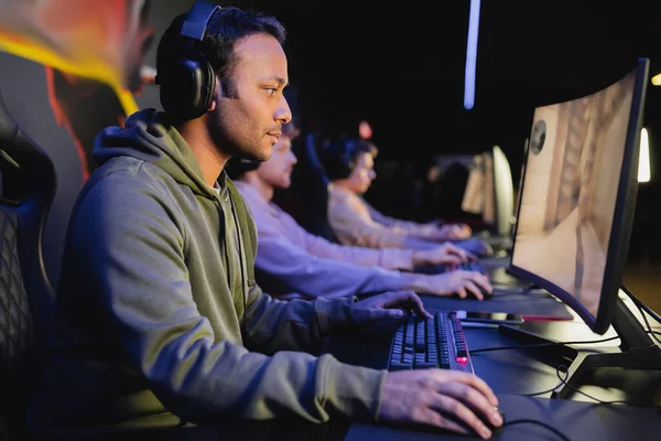 Side view of indian gamer in headphones playing video game with blurred team in club — Stock Photo