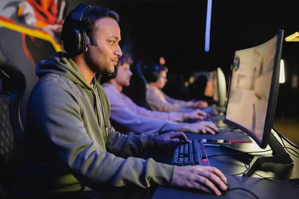 Side view of indian player in headphones playing video game with blurred team in cyber club — Stock Photo