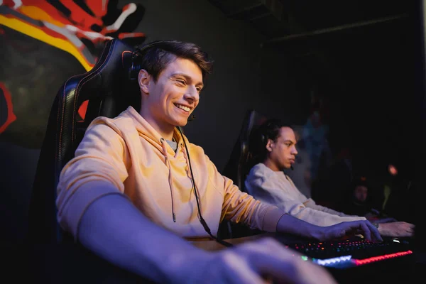 Cheerful gamer using keyboard near blurred multiracial friend in gaming club — Stock Photo