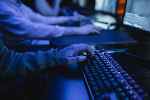 Обрезанный вид геймера с помощью клавиатуры рядом с монитором компьютера в игровом клубе с освещением — стоковое фото