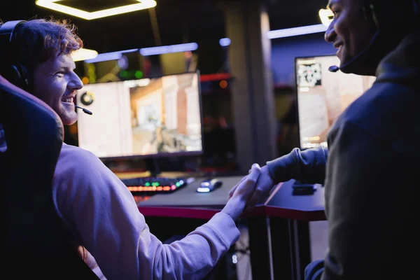 Des joueurs multiethniques souriants serrant la main près d'ordinateurs flous dans un club de jeu — Photo de stock