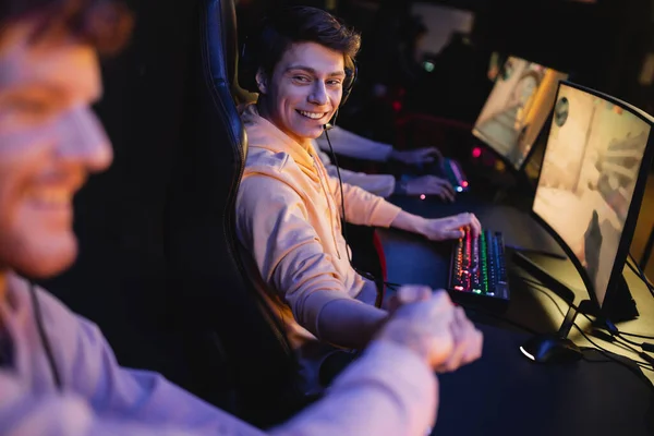 Hombre sonriente en auriculares estrechando la mano de un amigo borroso cerca de las computadoras en el club cibernético - foto de stock