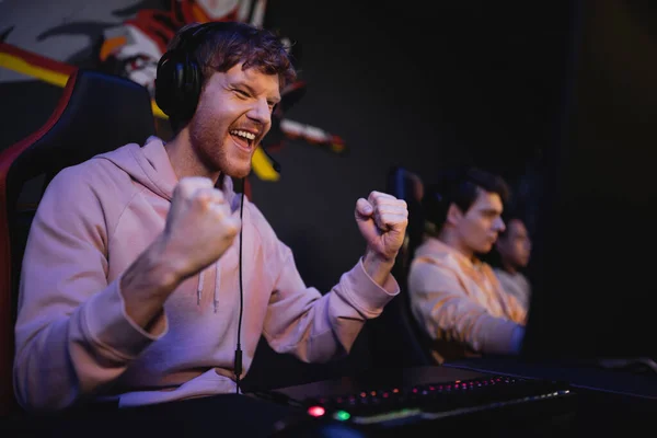 Возбужденный геймер в наушниках делает да жест рядом с размытой командой в игорном клубе — стоковое фото