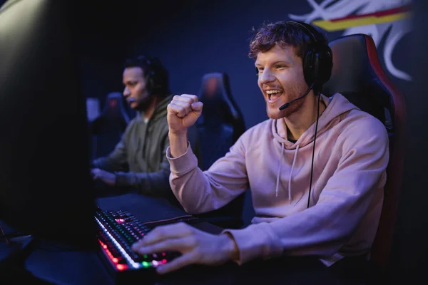 Возбужденный молодой геймер в наушниках показывает да жест рядом с компьютером в игровом клубе — стоковое фото