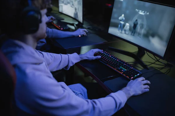 Jogador desfocado em fones de ouvido jogando jogo de computador no clube cibernético com iluminação — Fotografia de Stock