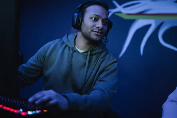 Indian gamer in headphones looking away near keyboard in cyber club — Stock Photo