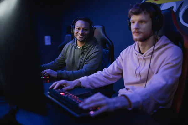 Smiling indian gamer in headphones looking at friend playing video game in cyber club — Stock Photo