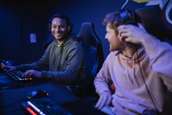 Smiling indian gamer in headphones sitting near blurred friend in cyber club — Stock Photo