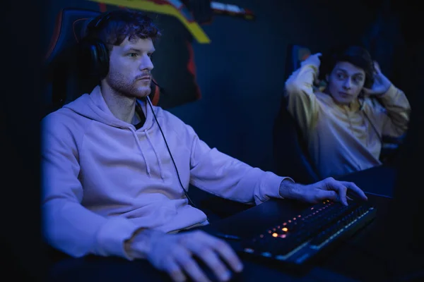Jogador focado em fones de ouvido usando teclado perto amigo borrado no clube cibernético — Fotografia de Stock