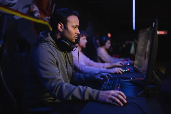 Side view of indian gamer playing video game on computer with blurred team in cyber club — Stock Photo