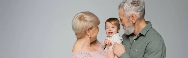 Mature woman with bearded husband looking at cheerful toddler granddaughter isolated on grey, banner — Stock Photo