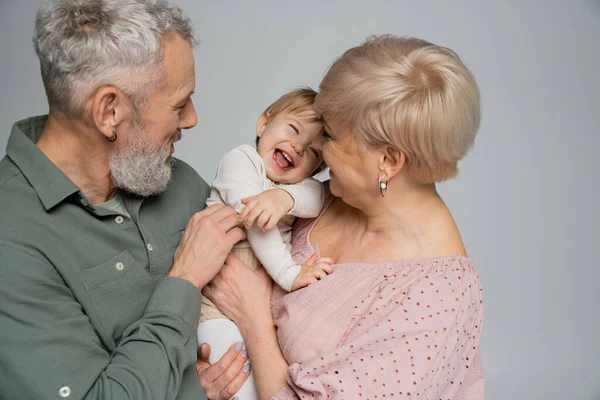 Excited toddler girl laughing near happy grandparents isolated on grey — Stock Photo
