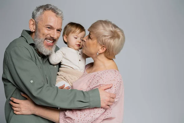 Mature woman kissing granddaughter near pleased bearded husband isolated on grey — Stock Photo