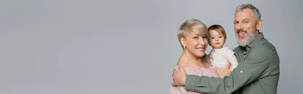 Joyful couple smiling at camera near little granddaughter isolated on grey, banner — Stock Photo
