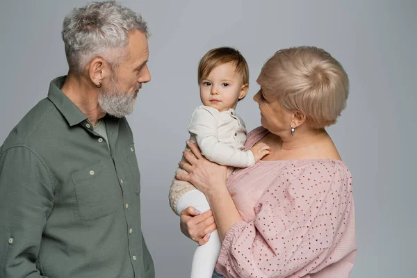 Senior femme tenant tout-petit petite-fille près barbu gris cheveux mari isolé sur gris — Photo de stock