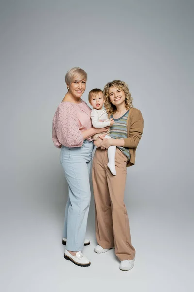 Полная длина зрелой женщины, улыбающейся в камеру рядом с взрослой дочерью с ребенком малыша на сером фоне — стоковое фото