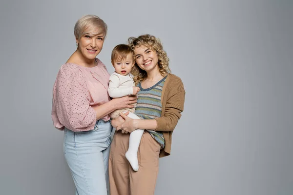 Pleased women embracing toddler girl and smiling at camera isolated on grey — Stock Photo