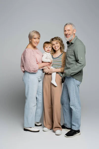 Longitud completa de los abuelos de mediana edad feliz y la hija adulta con la niña sonriendo a la cámara sobre fondo gris - foto de stock